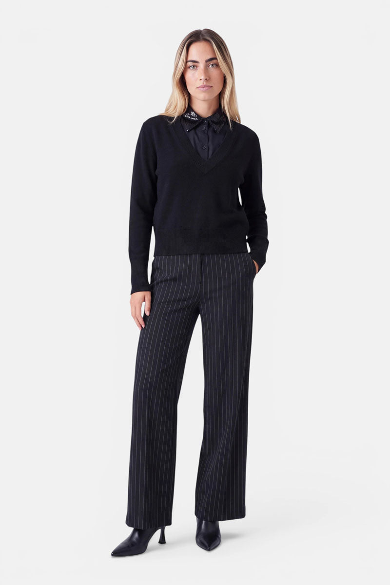 Eine Frau steht vor einem weißen Hintergrund und trägt einen schwarzen Pullover mit V-Ausschnitt und einem stilvollen Krageneinsatz über einer schwarzen Bluse im Blusen-Look mit dezenten Punkten, eine schwarze Nadelstreifenhose mit weitem Bein sowie schwarze Stiefel mit Absatz. Sie hat blondes Haar, einen neutralen Gesichtsausdruck und steckt ihre linke Hand in die Tasche.