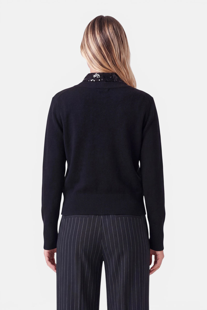 Vor einem schlichten weißen Hintergrund steht eine Person mit langen, blonden Haaren mit dem Rücken zur Kamera. Sie trägt einen schwarzen Langarmpullover mit einem Perlenbesatz am Kragen im stilvollen Blusen-Look sowie eine dunkelgraue Nadelstreifenhose.