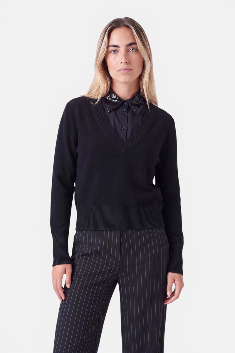 Eine Frau mit langen blonden Haaren trägt einen schwarzen Pullover mit V-Ausschnitt und dezentem Krageneinsatz, über einem schwarzen Hemd mit Kragen im Blusen-Look mit Verzierungen, dazu eine schwarze Nadelstreifenhose. Sie steht mit dem Gesicht nach vorne vor einem schlichten weißen Hintergrund und blickt mit neutralem Gesichtsausdruck in die Kamera.