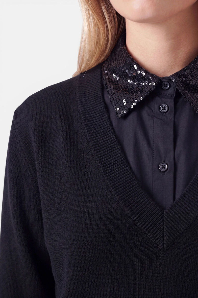 Eine Person trägt einen schwarzen Pullover mit V-Ausschnitt aus Baumwolle über einem schwarzen Hemd mit Knöpfen, das einen mit Pailletten verzierten Kragen und einen Krageneinsatz aufweist und so einen eleganten Blusen-Look erzeugt. Zu sehen sind nur der untere Teil des Gesichts, der Hals und der obere Brustbereich; die Person hat helle Haut und blonde Haare. Der Hintergrund ist einfarbig und hell.