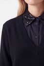 Eine Person trägt einen schwarzen Pullover mit V-Ausschnitt aus Baumwolle über einem schwarzen Hemd mit Knöpfen, das einen mit Pailletten verzierten Kragen und einen Krageneinsatz aufweist und so einen eleganten Blusen-Look erzeugt. Zu sehen sind nur der untere Teil des Gesichts, der Hals und der obere Brustbereich; die Person hat helle Haut und blonde Haare. Der Hintergrund ist einfarbig und hell.