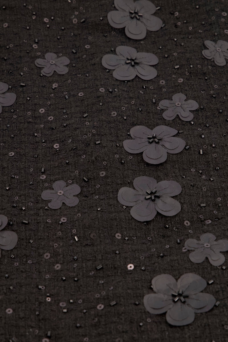 Ein dunkelgrauer Stoff in Tweed-Optik, verziert mit vereinzelten, reliefartigen 3D-Blumen. Der Stoff ist zudem mit kleinen, farblich passenden Pailletten und Perlen verziert.