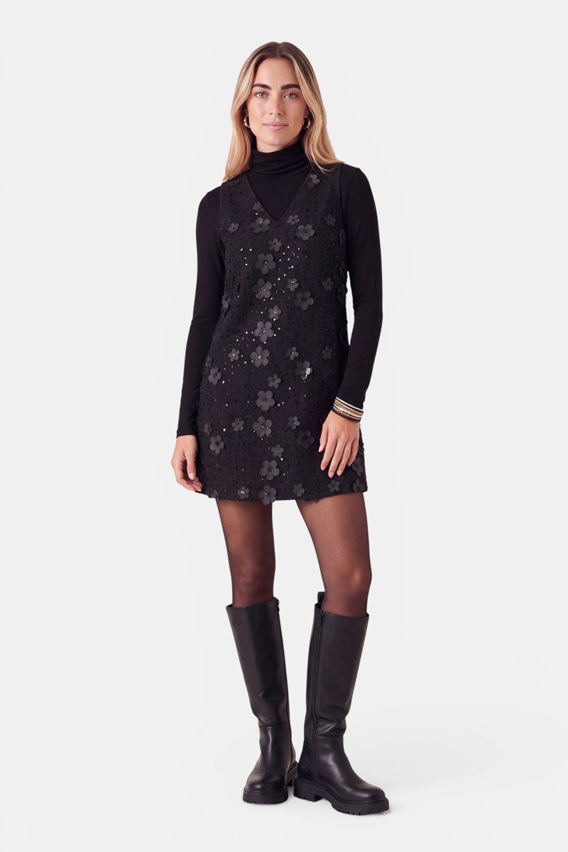 Eine Frau trägt ein schwarzes, ärmelloses Minikleid mit tweedartiger Optik, das mit 3D-Blumenapplikationen und Pailletten verziert ist, über einem schwarzen Rollkragenpullover. Dazu trägt sie eine transparente schwarze Strumpfhose und schwarze kniehohe Stiefel.