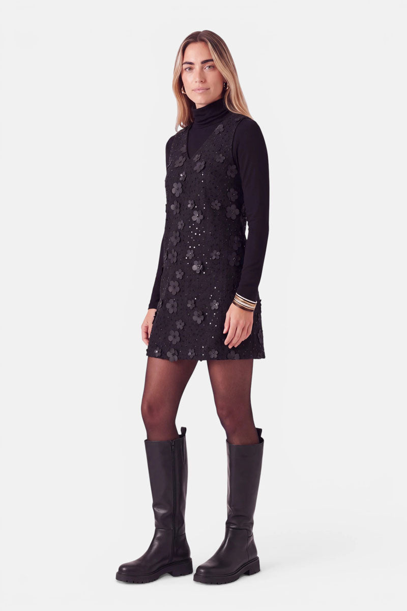 Eine Frau trägt ein schwarzes Minikleid, das mit 3D-Blumenapplikationen verziert ist, darüber einen schwarzen Rollkragenpullover und eine Strumpfhose, dazu schwarze kniehohe Stiefel.