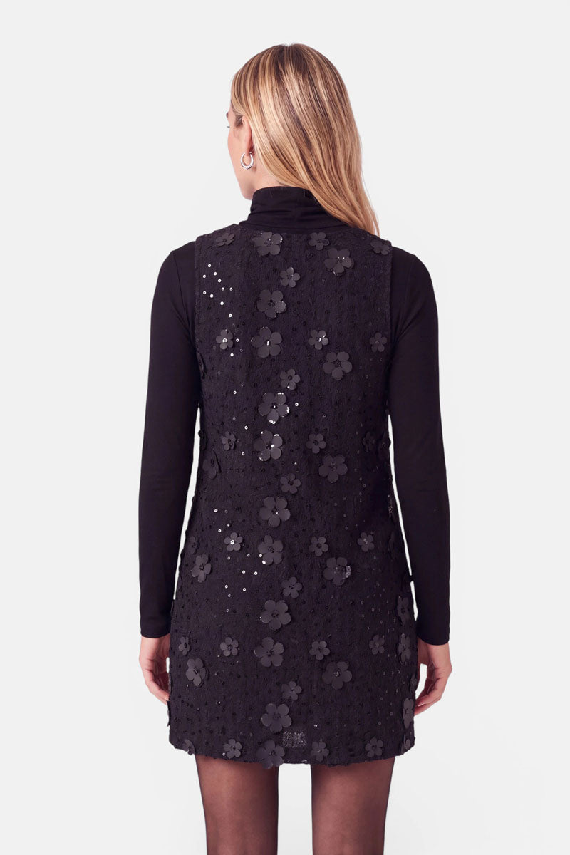 Eine Frau, die einen schwarzen, langärmeligen Rollkragenpullover unter einem ärmellosen, knielangen schwarzen Kleid im Tweed-Look trägt, das mit dreidimensionalen Blumenapplikationen und vereinzelten Pailletten verziert ist.