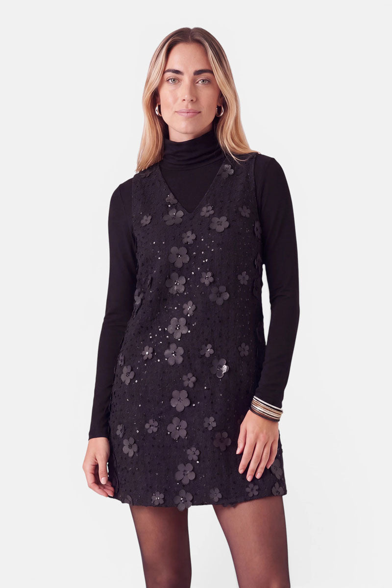Eine Frau trägt ein schwarzes, ärmelloses Minikleid mit 3D-Blumen und mit Pailletten verzierten Blumenapplikationen über einem schwarzen, langärmeligen Rollkragenpullover.