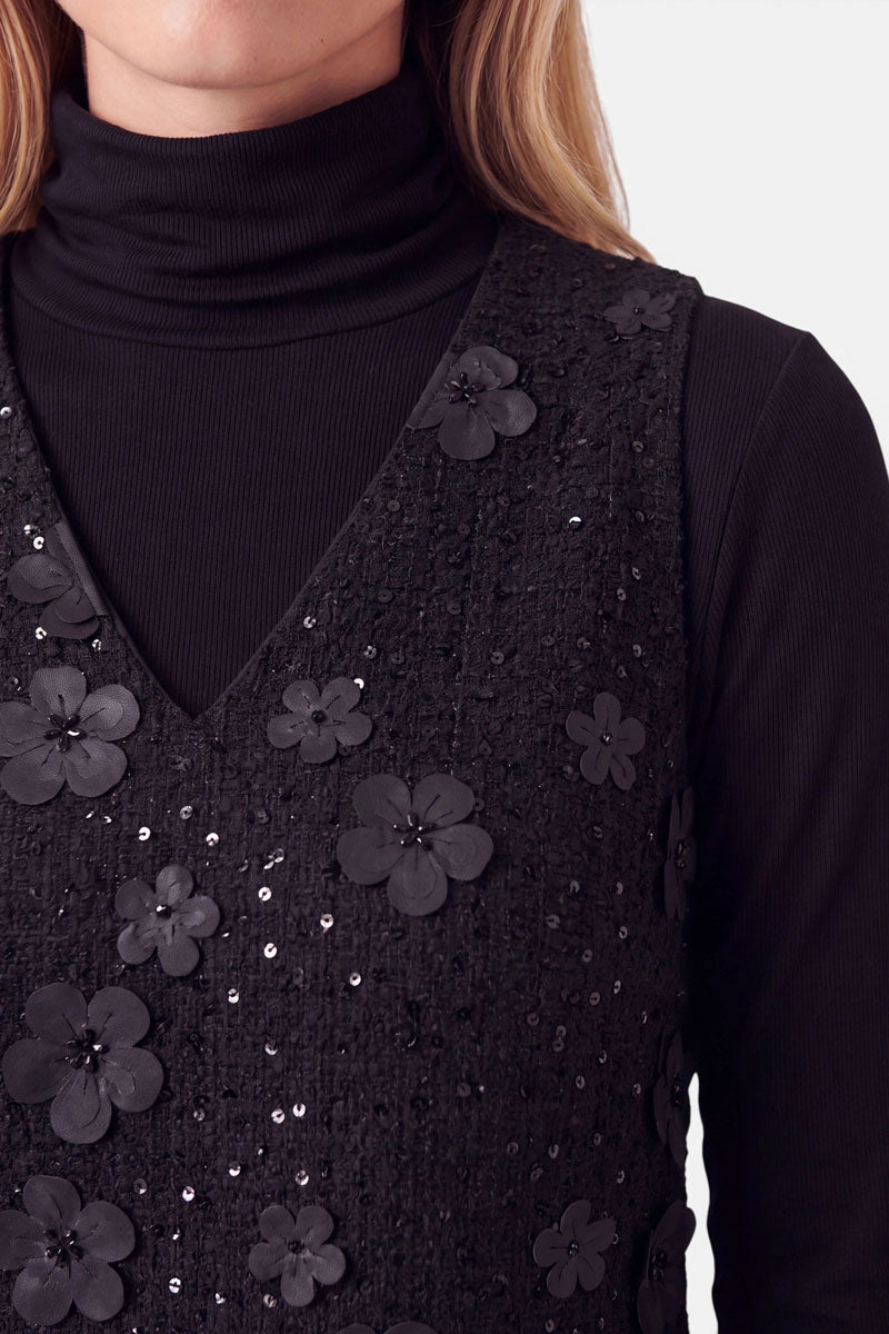 Eine Person trägt einen schwarzen Rollkragenpullover unter einem ärmellosen Minikleid mit V-Ausschnitt. Das Kleid ist mit vereinzelten Pailletten und dreidimensionalen Stoffblumen verziert.