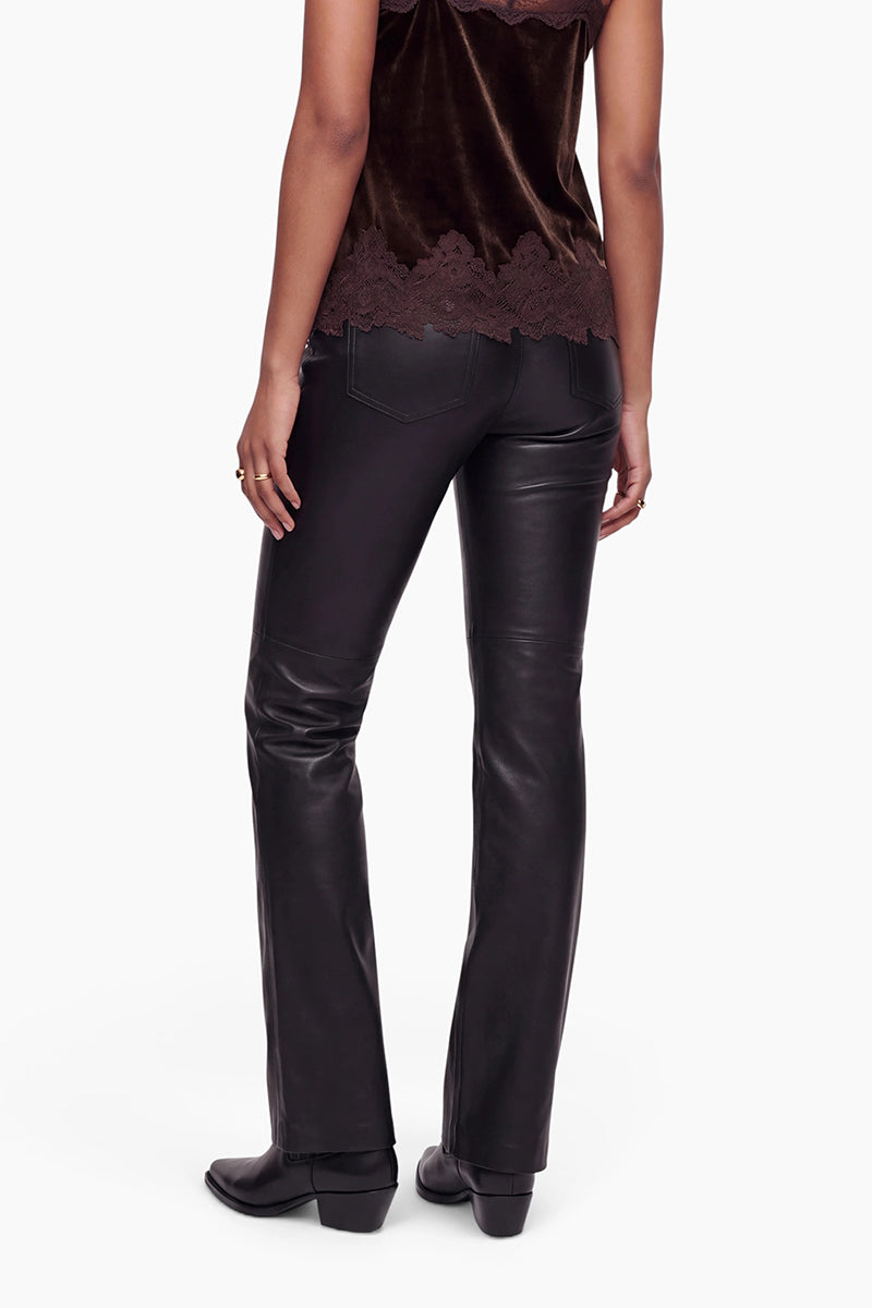 Eine Person steht mit dem Rücken zur Kamera und trägt eine schwarze vegane Lederhose, dazu schwarze Schuhe mit Absatz und ein dunkelbraunes Samtop mit Spitzenbesatz am Saum. Der Hintergrund ist schlicht weiß. Zu sehen sind nur der untere Teil des Oberkörpers, die Beine und ein Teil der Arme der Person.