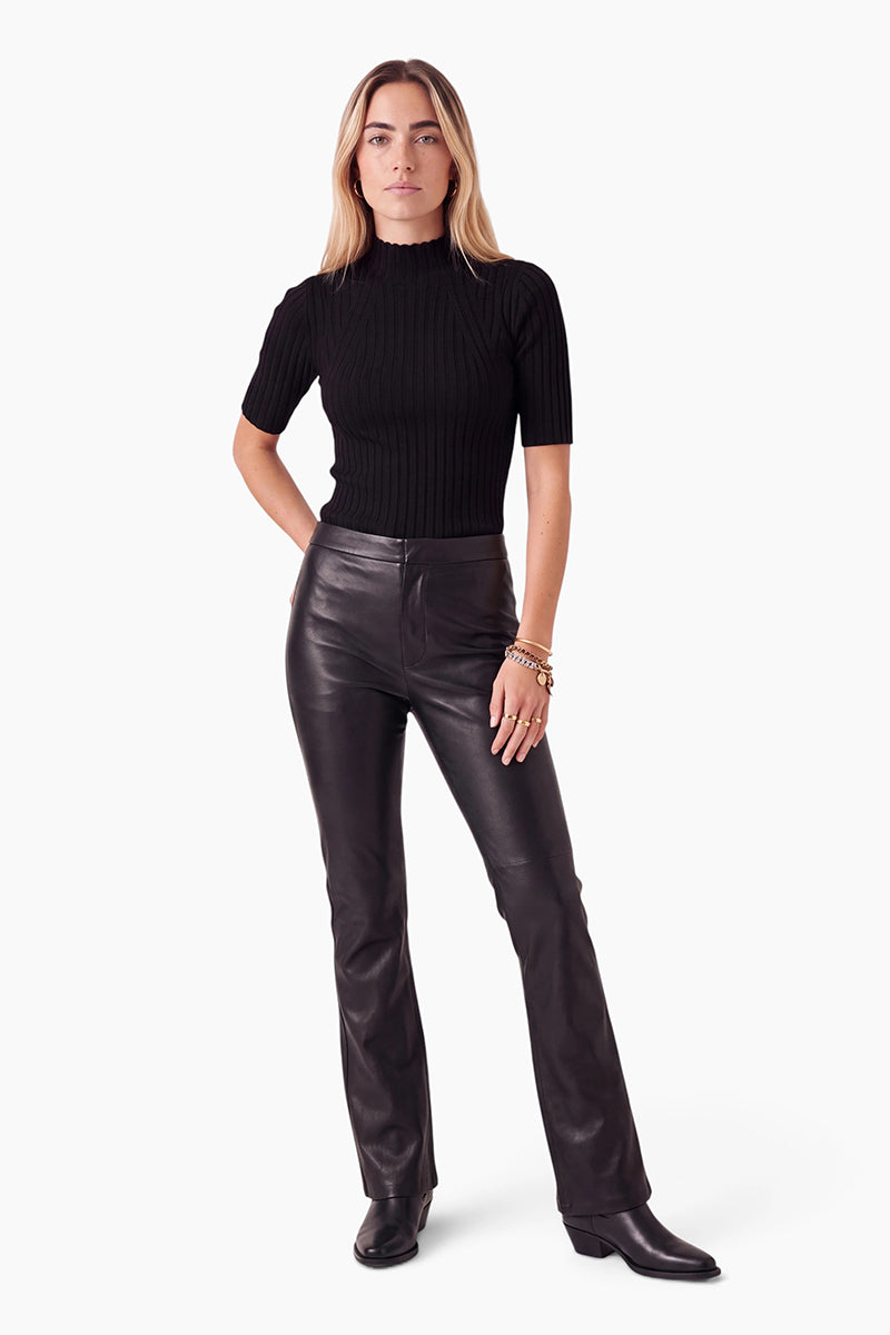 Eine Frau steht mit dem Blick nach vorne und trägt ein eng anliegendes schwarzes, geripptes Kurzarm-Oberteil, eine hoch taillierte vegane Lederhose in gerader Passform sowie schwarze Stiefel mit Absatz. Sie hat langes, glattes blondes Haar und trägt nur wenig Schmuck. Der Hintergrund ist schlicht weiß.