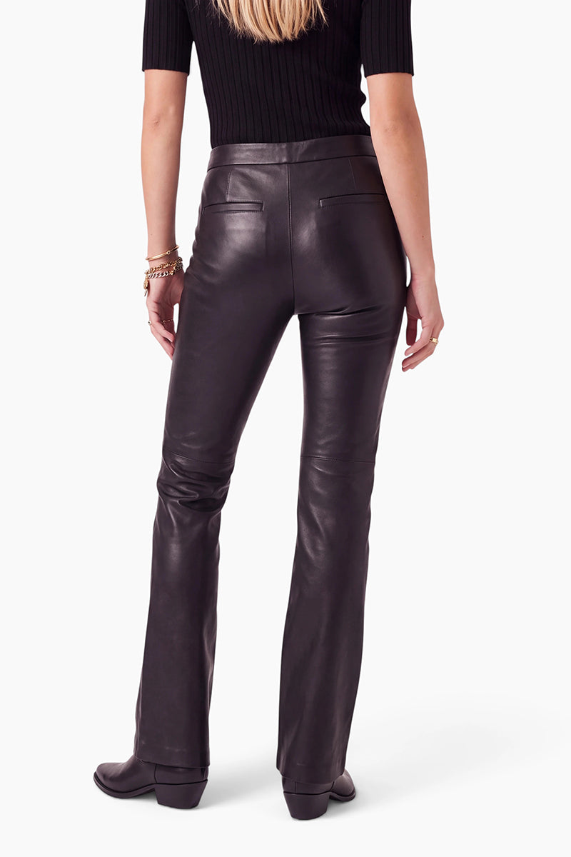 Eine Person ist von hinten zu sehen. Sie trägt ein schwarzes, kurzärmeliges, geripptes Oberteil und eine hoch taillierte, figurbetonte vegane Lederhose mit zwei Gesäßtaschen. Sie hat langes blondes Haar, schwarze Stiefel und Armbänder an einem Handgelenk. Der Hintergrund ist einfarbig weiß.