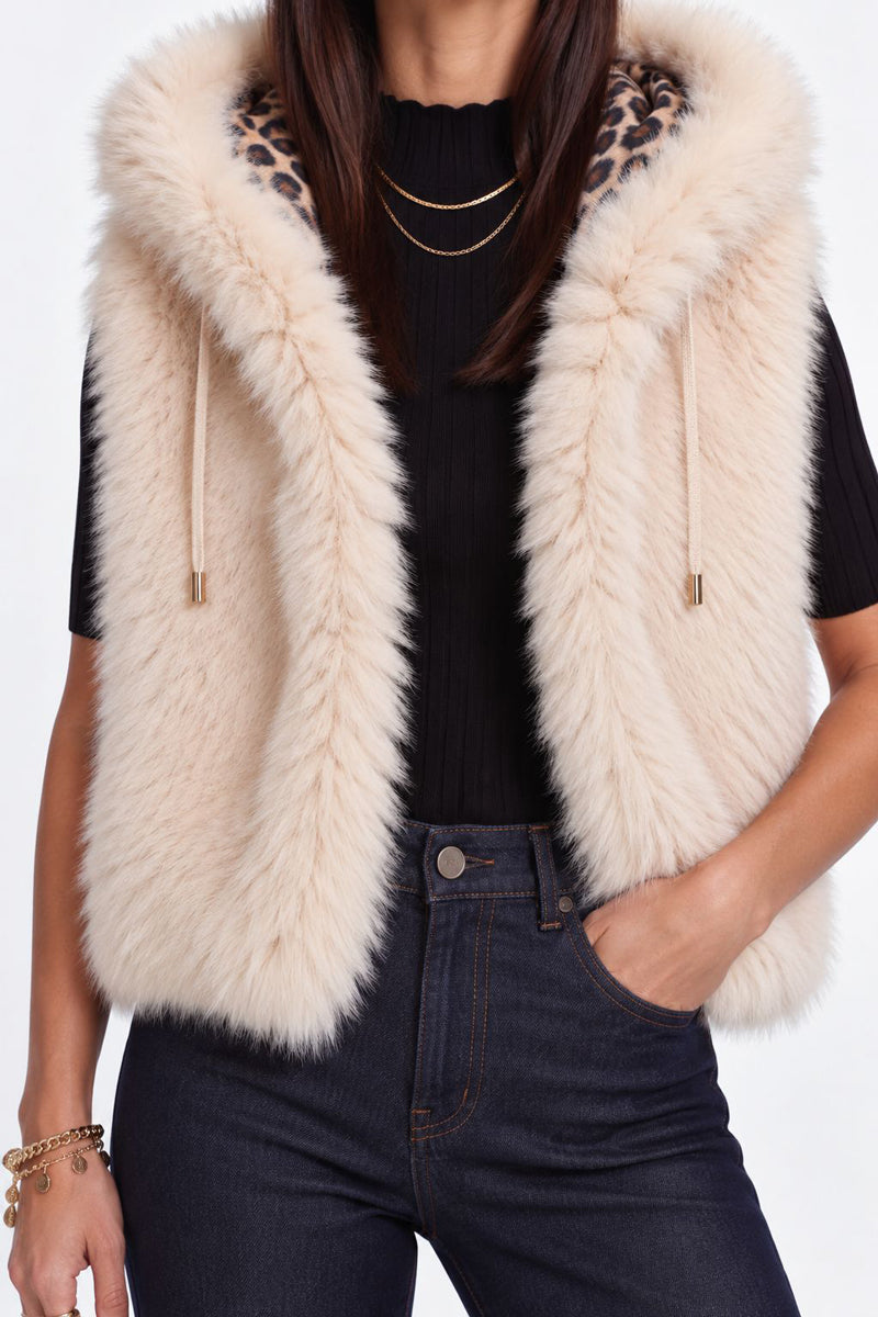Eine Frau trägt eine ärmellose, beige Kunstfellweste mit Kapuze, deren Kapuze luxuriös mit Leo-Futter gefüttert ist, über einem schwarzen, gerippten Oberteil und einer dunklen Jeans mit hoher Taille. Als Accessoires trägt sie mehrere goldene Halsketten und ein Armband; eine Hand steckt in ihrer Jeanstasche.