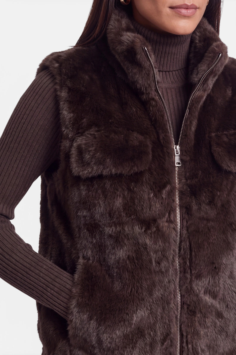 Eine Frau trägt eine dunkelbraune Kunstfellweste mit Reißverschluss über einem gerippten, langärmeligen Rollkragenpullover. Ihre linke Hand steckt in einer Westentasche. Das Bild ist an der Nase und auf halber Armhöhe beschnitten und vor einem schlichten weißen Hintergrund aufgenommen.