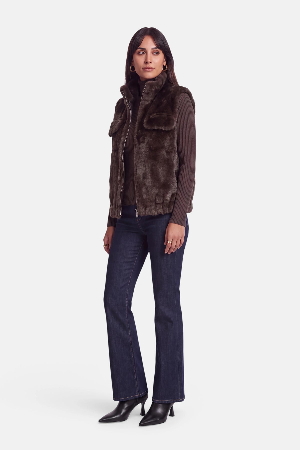 Eine Frau steht in einer Dreiviertelansicht und trägt eine braune Kunstfellweste mit Reißverschluss über einem gerippten braunen Rollkragenpullover mit Stehkragen, dunkelblaue Jeans und schwarze Stiefel mit Absatz. Sie hat dunkles Haar, trägt nur wenig Schmuck und hat einen neutralen Gesichtsausdruck, vor einem schlichten weißen Hintergrund.