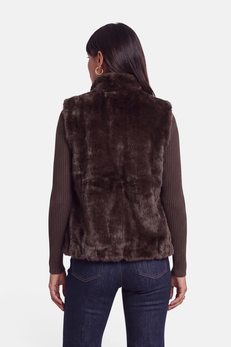 Eine Frau, die von hinten zu sehen ist, trägt eine dunkelbraune Kunstfellweste mit Stehkragen über einem gerippten, langärmeligen Oberteil und einer dunklen Jeans. Sie hat glattes, langes dunkles Haar und goldene Creolen. Der Hintergrund ist einfarbig weiß.