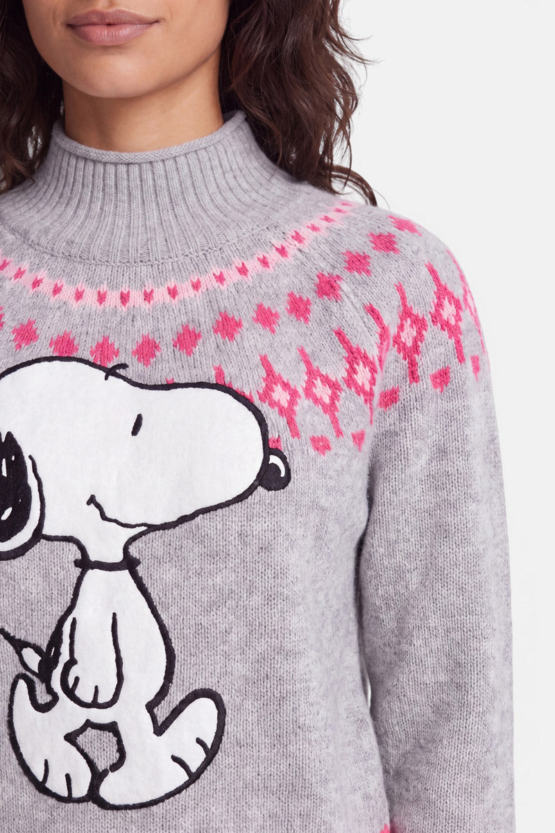 Eine Frau mit welligem braunem Haar trägt einen grauen Pullover aus Merinowolle mit einem großen Snoopy-Aufdruck und einem rosa Rautenmuster im norwegischen Stil auf der Brust. Der Pullover hat einen gerippten Rollkragen. Ihr Gesicht ist teilweise zu sehen, man erkennt ihre Lippen und ihr Kinn.