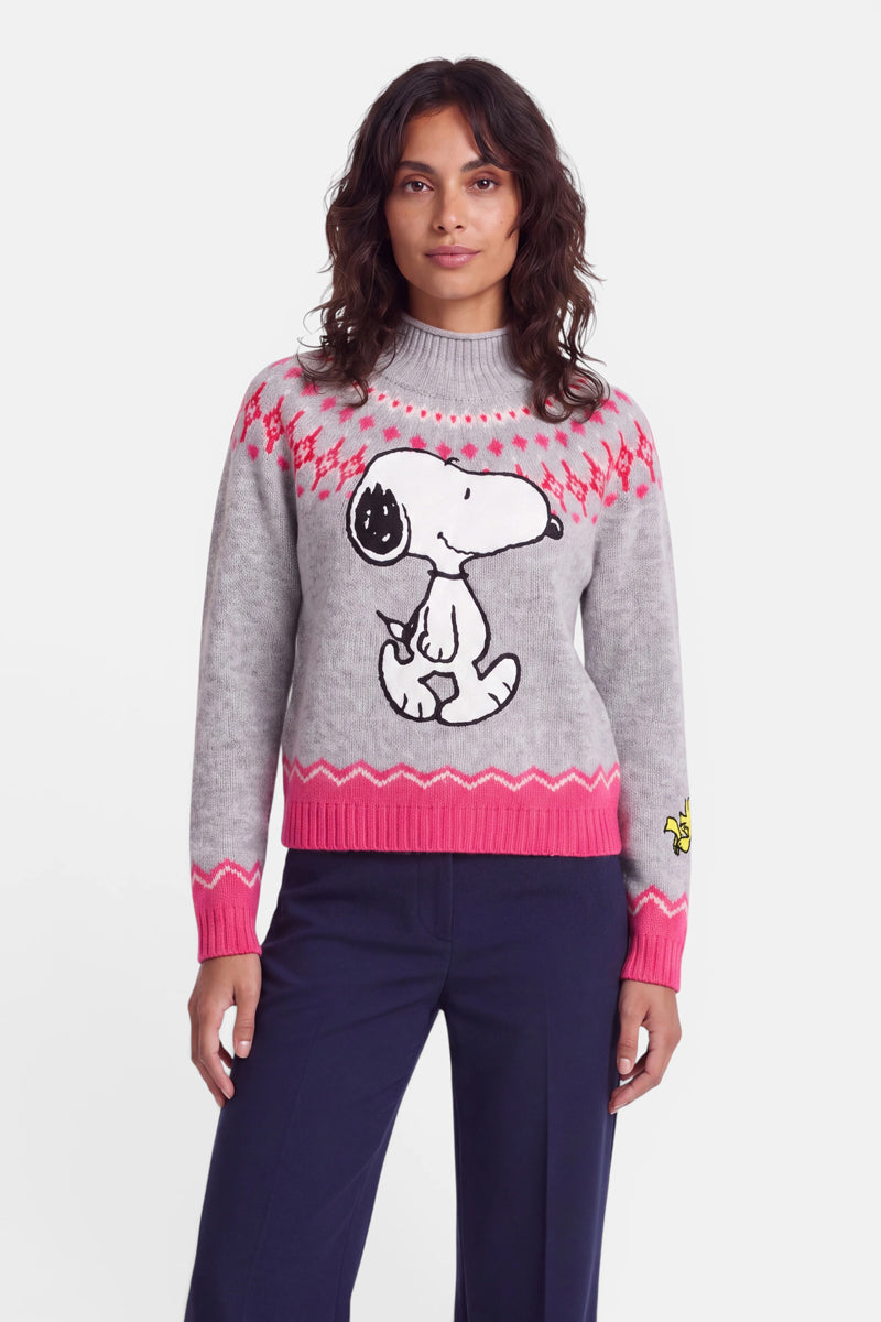 Eine Frau trägt einen grauen Pullover im Norweger-Stil mit rosa Mustern, auf dessen Vorderseite ein großes Snoopy-Motiv und auf dem Ärmel Woodstock abgebildet ist. Der Pullover aus Merinowolle hat einen hohen Kragen und rosa Bündchen; dazu trägt sie eine dunkelblaue Hose. Sie steht mit dem Gesicht nach vorne vor einem weißen Hintergrund.
