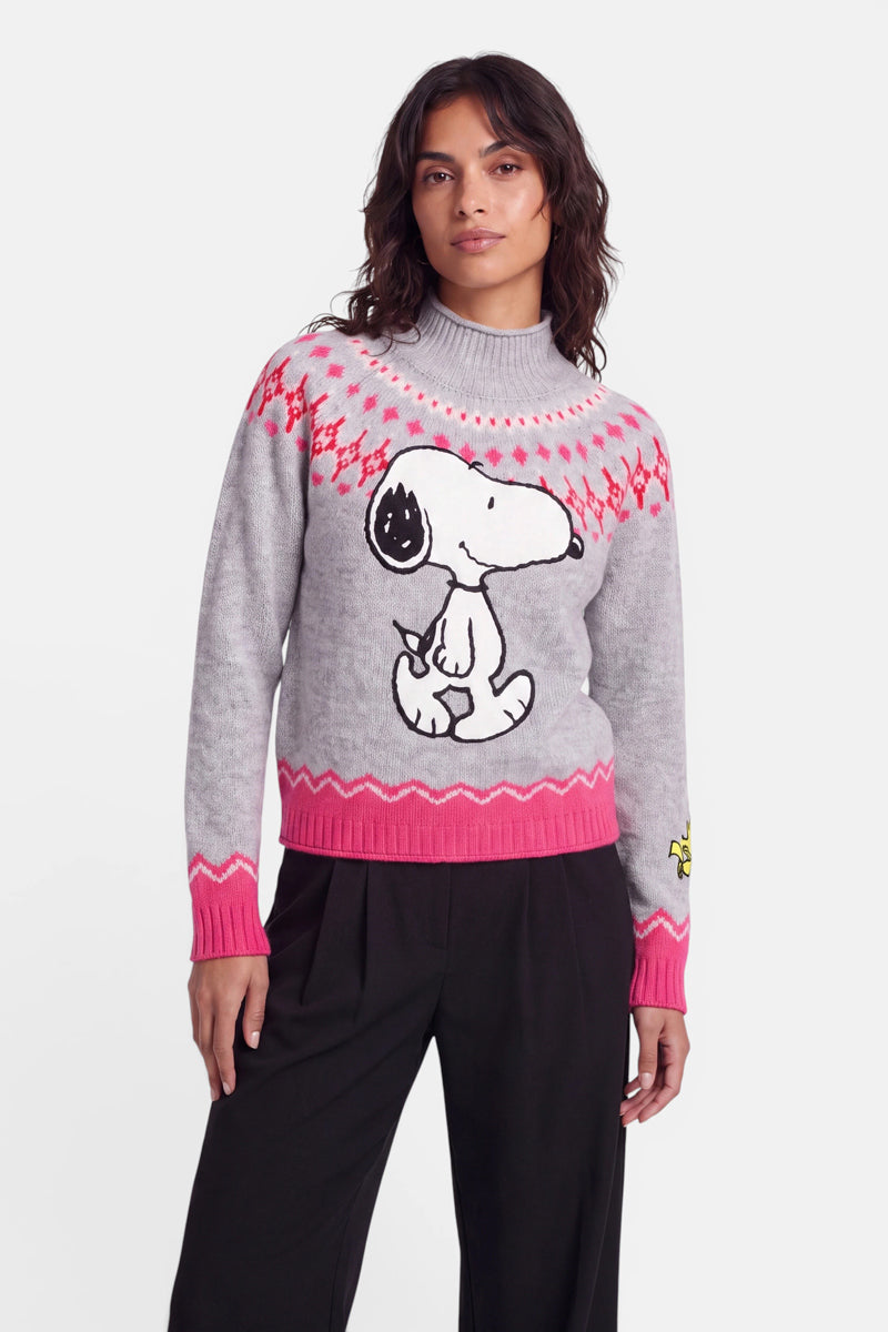 Eine Frau trägt einen hellgrauen Pullover im norwegischen Stil mit einem großen Snoopy-Aufdruck, einem rosa-roten Muster im Schulterbereich, rosa Bündchen und Saum sowie eine schwarze Hose. Der Pullover aus weicher Merinowolle ist außerdem auf dem linken Ärmel mit einer kleinen gelben Woodstock-Figur verziert. Sie steht vor einem schlichten weißen Hintergrund.