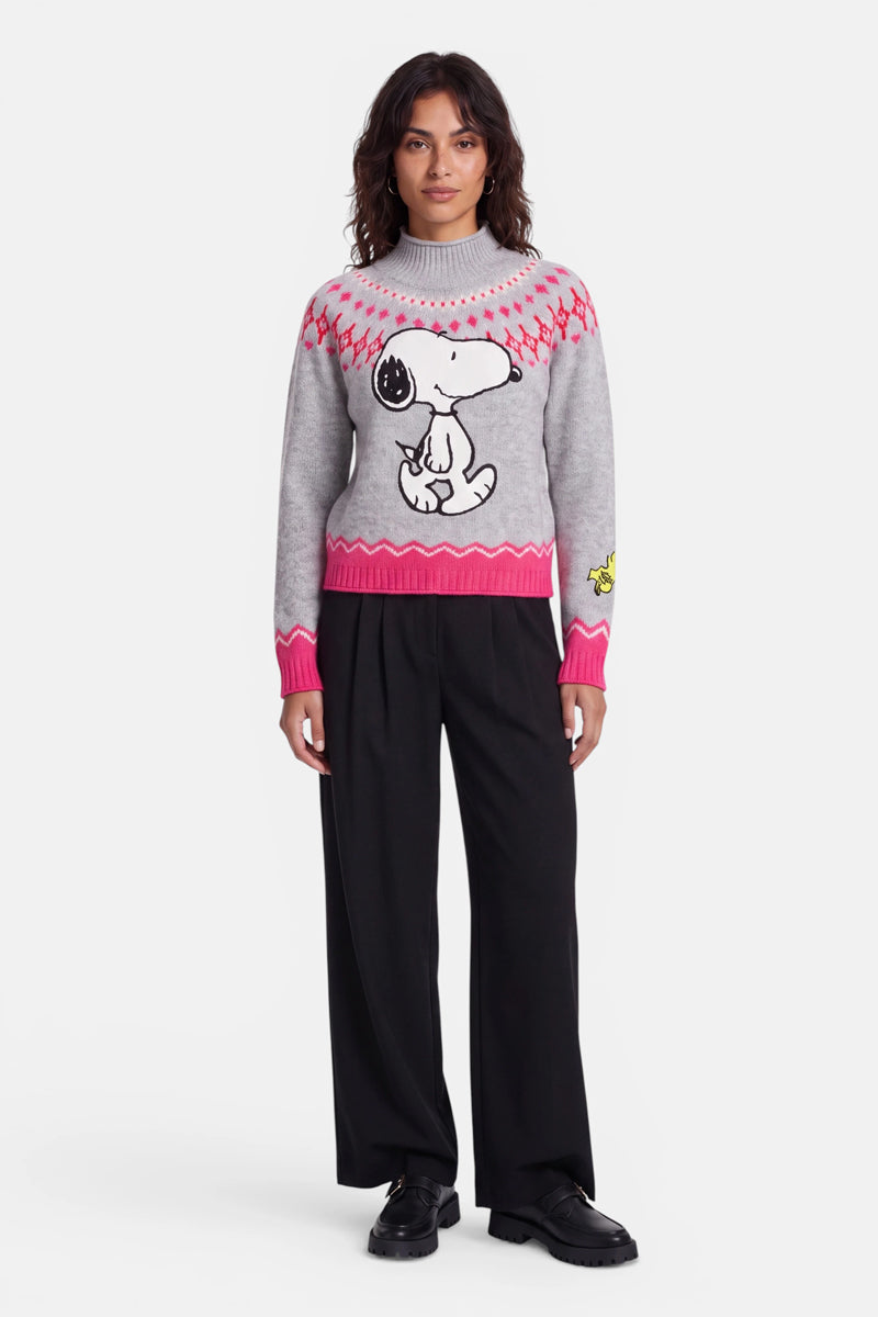 Eine Person steht mit dem Gesicht nach vorne und trägt einen grauen Pullover im norwegischen Stil mit rosa Mustern, auf dem ein großes Snoopy-Motiv und ein kleiner Woodstock auf dem Ärmel abgebildet sind. Sie hat dunkles, welliges Haar, trägt eine schwarze Hose mit weitem Bein und schwarze Schuhe vor einem weißen Hintergrund.