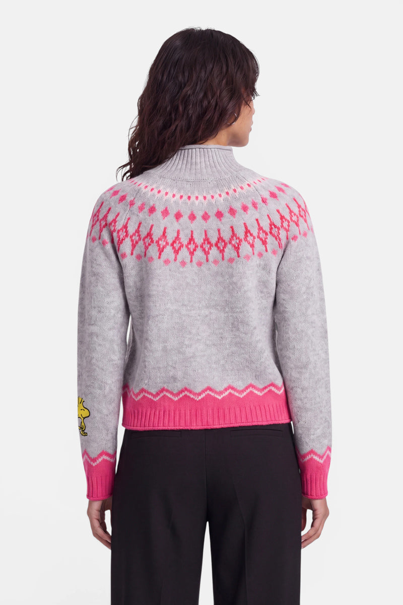 Eine Person mit welligem, dunklem Haar trägt einen hellgrauen Pullover im norwegischen Stil, der an den Schultern und an den Ärmelbündchen mit einem rosa geometrischen Muster verziert ist und auf dem linken Ärmel eine kleine gelbe Comicfigur zeigt. Der Pullover hat einen hohen Kragen und wird zu einer schwarzen Hose getragen. Die Person ist mit dem Rücken zur Kamera gewandt.
