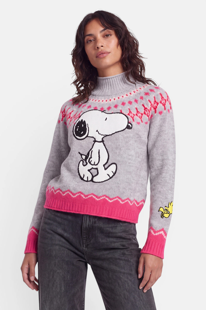 Eine Person trägt einen grauen, hochgeschlossenen Pullover aus Merinowolle mit rosa Akzenten im norwegischen Stil. Auf der Vorderseite ist ein großes Snoopy-Motiv zu sehen, auf dem Ärmel ein kleines Woodstock-Motiv. Der Pullover wird zu einer dunklen Jeans kombiniert, und die Person steht vor einem schlichten weißen Hintergrund.