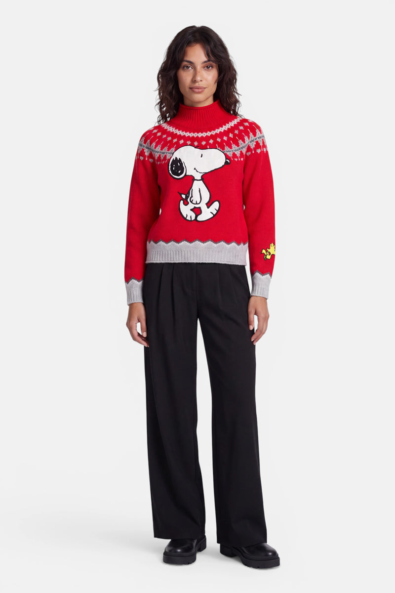 Eine Person steht mit dem Gesicht nach vorne und trägt einen roten Pullover mit Snoopy-Motiv, auf dem Snoopy und Woodstock aus „Peanuts“ abgebildet sind. Der Pullover hat graue Bündchen und einen grauen Saum mit dezentem norwegischem Muster und wird zu einer schwarzen Hose mit weitem Bein und schwarzen Schuhen kombiniert. Der Hintergrund ist einfarbig weiß.