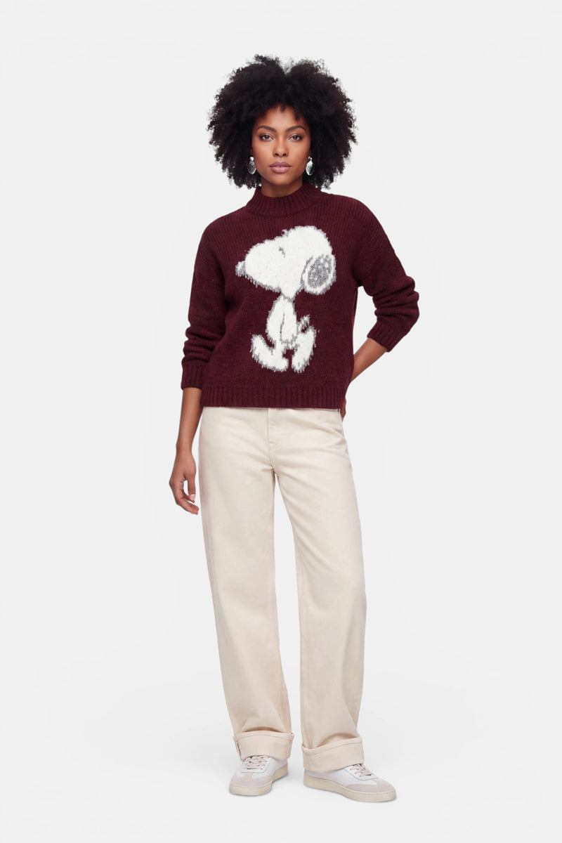Eine Frau steht mit dem Blick nach vorne und trägt einen burgunderroten Grobstrickpullover mit einem großen Snoopy-Motiv auf der Vorderseite, dazu eine cremefarbene Hose mit weitem Bein und weiße Sneaker. Sie hat mittel-dunkle Haut, natürlich lockiges Haar und einen neutralen Gesichtsausdruck. Der Hintergrund ist schlicht weiß.