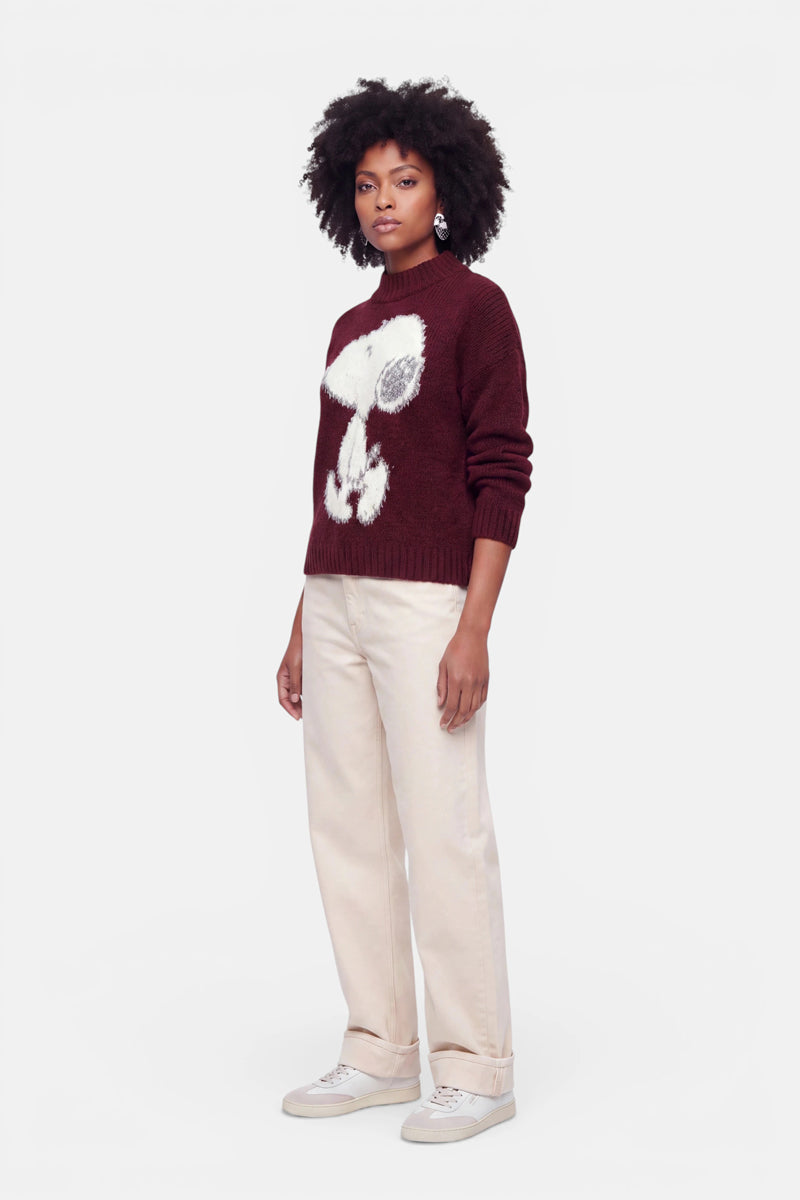 Eine Frau mit natürlich lockigem Haar steht mit dem Blick nach vorne. Sie trägt einen burgunderroten Grobstrick-Pullover mit einem großen weißen Snoopy-Motiv, eine cremefarbene Hose mit weitem Bein und weiße Sneaker. Der Hintergrund ist schlicht weiß. Ihr Gesichtsausdruck ist neutral, und sie trägt nur wenige Accessoires.