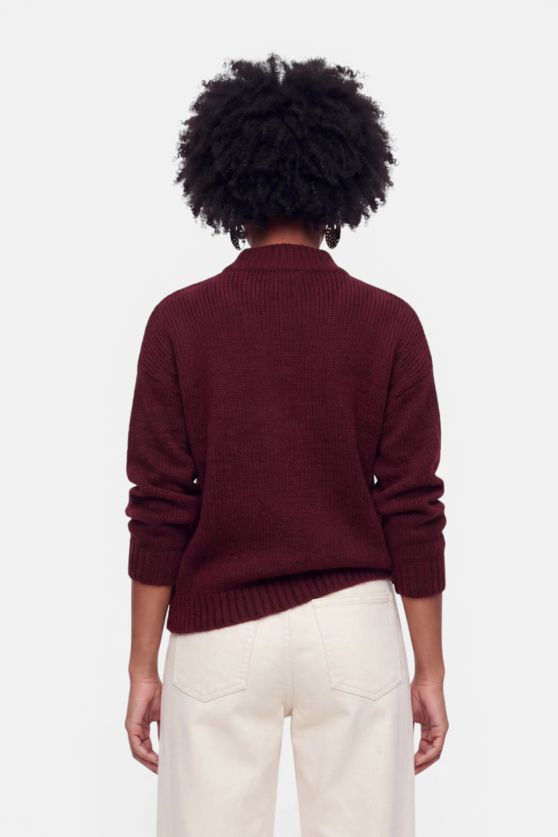 Eine Person steht mit dem Rücken zur Kamera und trägt einen kastanienbraunen Grobstrickpullover aus einer Woll-Mohair-Mischung mit langen Ärmeln sowie eine cremefarbene Hose mit hoher Taille. Die Arme hängen entspannt an den Seiten herab. Die Person hat kurzes, lockiges dunkles Haar. Der Hintergrund ist einfarbig und hell.