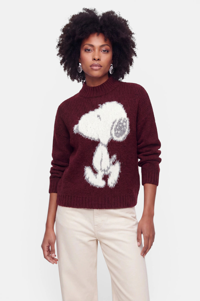 Eine Frau mit dunklem, lockigem Haar trägt hängende Ohrringe und einen burgunderroten Grobstrickpullover mit einem großen weißen Snoopy-Motiv. Der kuschelige Pullover aus einer Woll-Mohair-Mischung wird zu einer cremefarbenen Hose kombiniert. Sie steht vor einem schlichten hellgrauen Hintergrund, blickt mit ausdruckslosem Gesicht geradeaus.