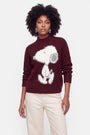 Eine Frau mit dunklem, lockigem Haar trägt hängende Ohrringe und einen burgunderroten Grobstrickpullover mit einem großen weißen Snoopy-Motiv. Der kuschelige Pullover aus einer Woll-Mohair-Mischung wird zu einer cremefarbenen Hose kombiniert. Sie steht vor einem schlichten hellgrauen Hintergrund, blickt mit ausdruckslosem Gesicht geradeaus.