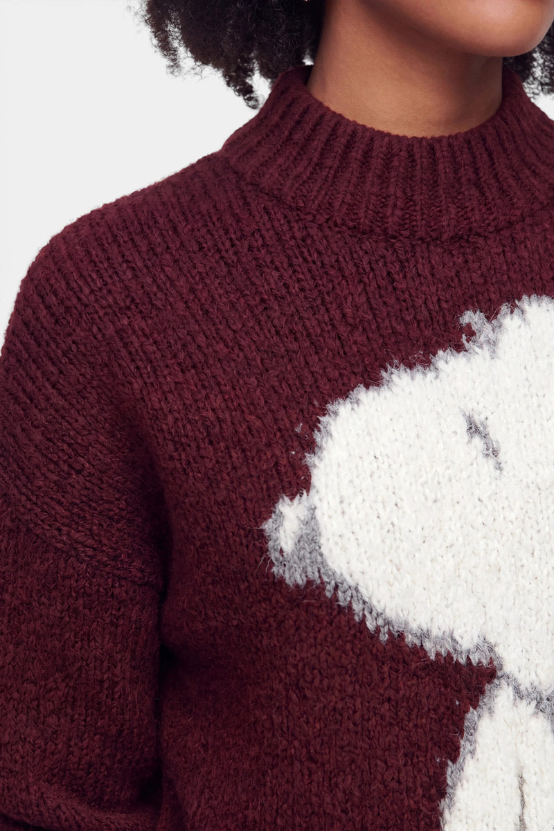 Nahaufnahme einer Person, die einen burgunderroten Grobstrickpullover mit hohem Kragen trägt. Der Pullover aus einer weichen Wolle-Mohair-Mischung ist auf der Vorderseite mit einem großen, flauschigen weißen Snoopy-Motiv verziert, das teilweise zu sehen ist. Zu sehen sind nur der untere Teil des Gesichts und der Hals der Person.