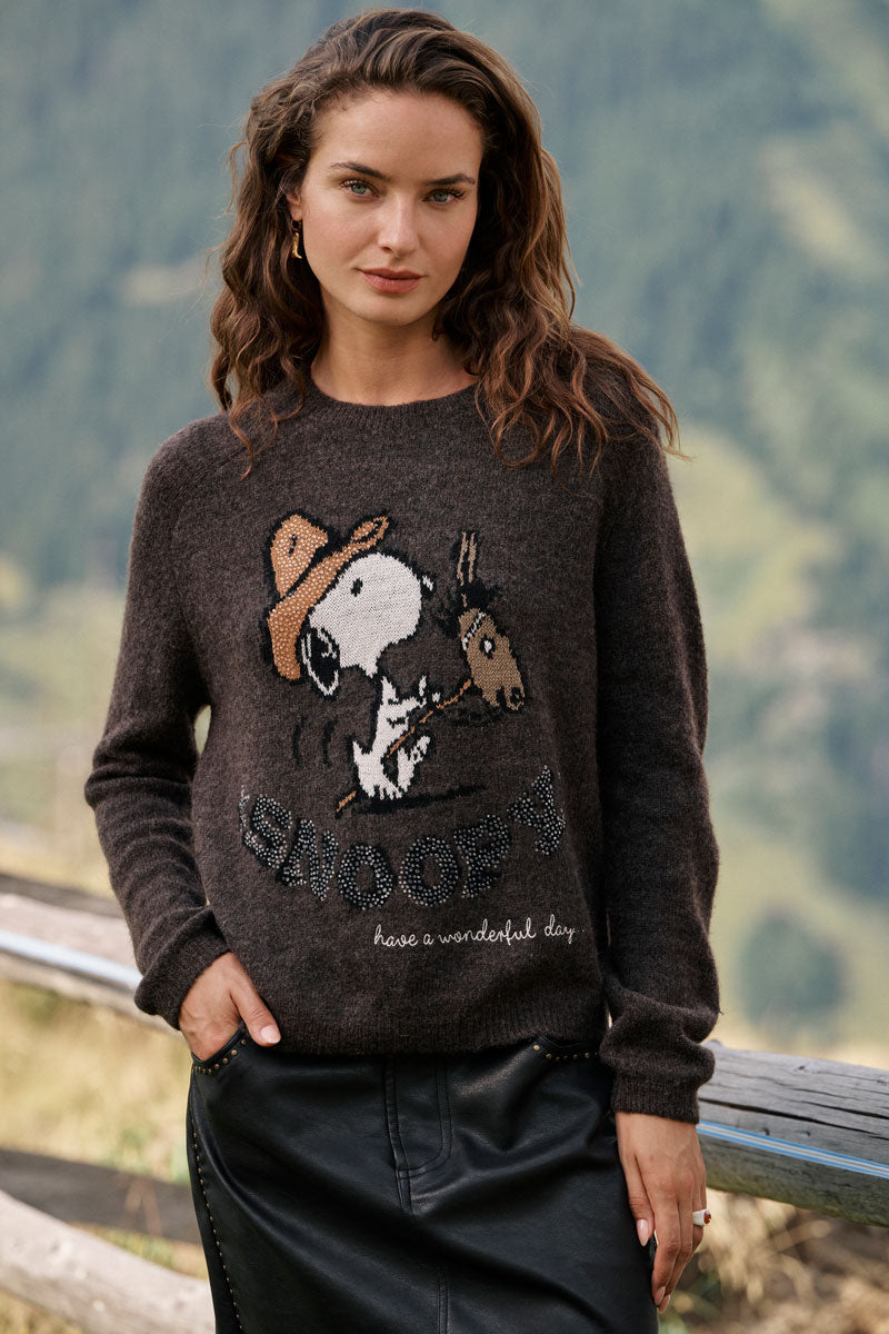 Eine Frau mit welligem braunem Haar trägt einen dunklen Snoopy-Intarsia-Pullover, auf dem Snoopy und Woodstock abgebildet sind, zusammen mit dem Schriftzug „Peanuts“ und „Have a wonderful day“. Sie steht draußen neben einem Holzgeländer, im Hintergrund sind verschwommene grüne Berge und Bäume zu sehen.