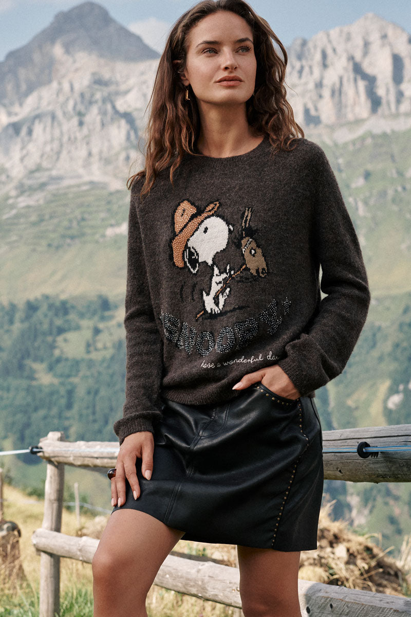 Eine Frau steht im Freien neben einem Holzzaun. Sie trägt einen dunklen Pullover mit einem Aufdruck von Snoopy und einem Pferd sowie einen kurzen schwarzen Minirock aus Kunstleder mit Gürtelschlaufen und Nietenverzierung. Hinter ihr erstrecken sich grüne Hügel und hohe Berge unter einem klaren Himmel. Sie hat langes braunes Haar und einen entspannten Gesichtsausdruck.