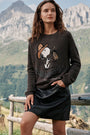 Eine Frau steht im Freien in der Nähe eines Holzzauns, im Hintergrund sind Berge zu sehen. Sie trägt einen braunen Pullover aus Alpaka-Wolle mit einem niedlichen Snoopy-Intarsienmuster und einem Pferd, dazu einen schwarzen Lederrock mit Strassverzierung. Ihre Hand ruht auf dem Zaun, und sie hat langes, braunes, welliges Haar.