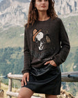 Eine Frau steht im Freien in der Nähe eines Holzzauns, im Hintergrund sind Berge zu sehen. Sie trägt einen braunen Pullover aus Alpaka-Wolle mit einem niedlichen Snoopy-Intarsienmuster und einem Pferd, dazu einen schwarzen Lederrock mit Strassverzierung. Ihre Hand ruht auf dem Zaun, und sie hat langes, braunes, welliges Haar.