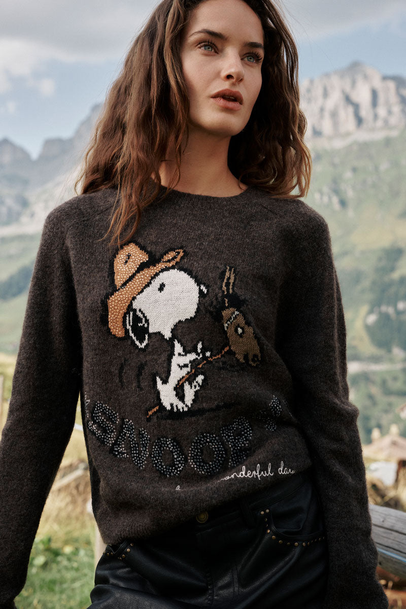 Eine Frau mit welligem braunem Haar steht im Freien in einer bergigen Landschaft und trägt einen dunklen Snoopy-Intarsia-Pullover mit einem niedlichen Motiv von Snoopy und Woodstock. Sie hat die Hände in den Taschen ihrer schwarzen Hose und blickt mit entspanntem Gesichtsausdruck in die Ferne.