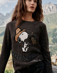 Eine Frau mit welligem braunem Haar steht im Freien in einer bergigen Landschaft und trägt einen dunklen Snoopy-Intarsia-Pullover mit einem niedlichen Motiv von Snoopy und Woodstock. Sie hat die Hände in den Taschen ihrer schwarzen Hose und blickt mit entspanntem Gesichtsausdruck in die Ferne.