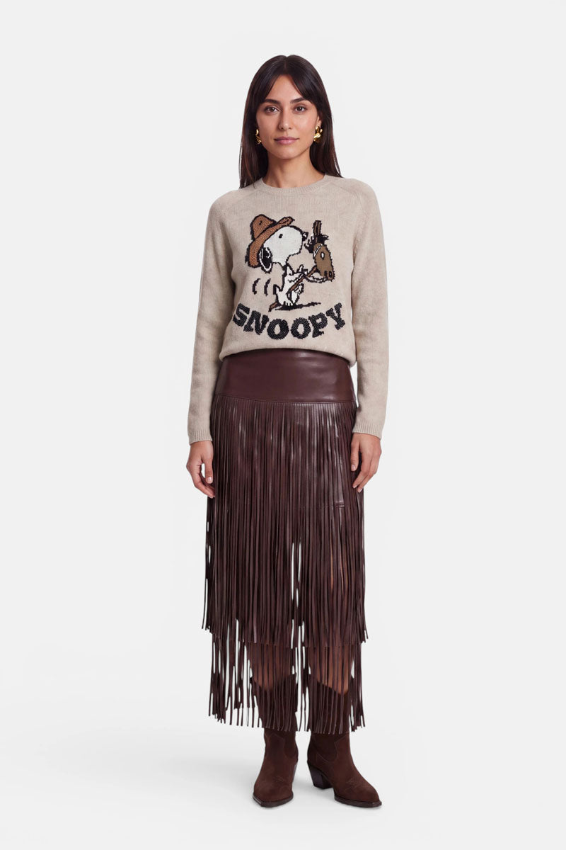 Eine Frau steht mit dem Blick nach vorne und trägt einen beigen Pullover mit einem Snoopy- und Woodstock-Motiv, einen braunen, mit Fransen verzierten veganen Lederminirock sowie braune Wildlederstiefel. Sie hat langes braunes Haar, eine entspannte Körperhaltung und einen neutralen Gesichtsausdruck und steht vor einem schlichten weißen Hintergrund.