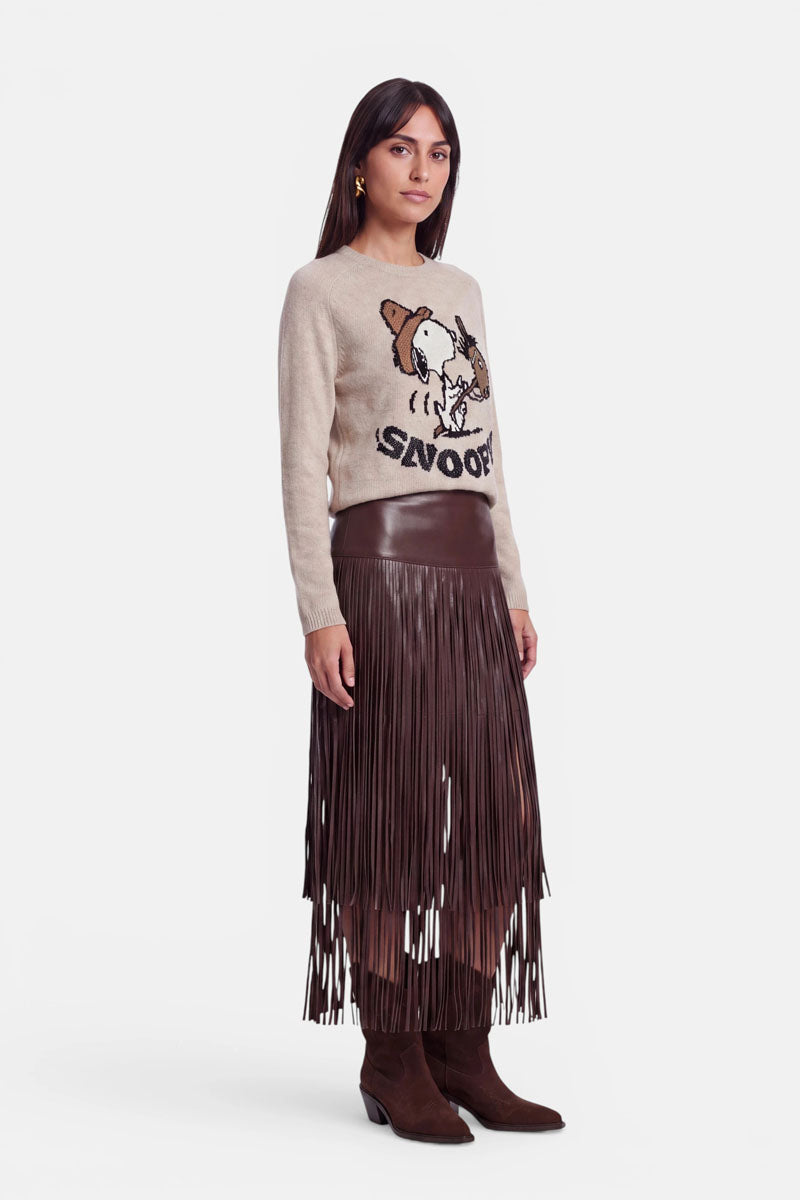 Eine Frau steht da und trägt einen beigen Pullover mit einem Snoopy-Motiv, einen braunen Minirock aus veganem Leder mit langen Fransen sowie braune Wildlederstiefel. Sie hat glattes dunkles Haar, goldene Ohrringe und einen neutralen Gesichtsausdruck und posiert vor einem schlichten weißen Hintergrund.