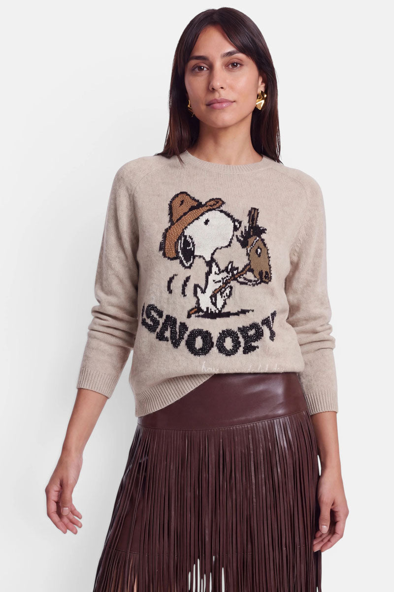 Eine Frau trägt einen hellbraunen Pullover aus Alpaka-Wolle, auf dem Snoopy mit einer Mütze und der Aufschrift „Snoopy!“ in verspielter Intarsienstickerei abgebildet ist. Dazu trägt sie einen braunen Lederrock mit Fransen. Ihr Haar ist offen, und sie trägt goldene Ohrringe. Der Hintergrund ist schlicht weiß.