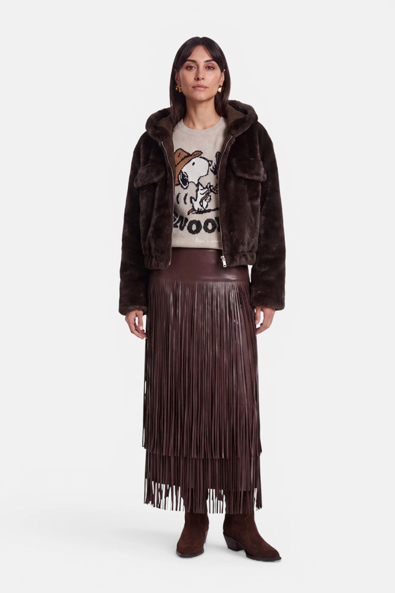 Eine Frau steht mit dem Blick nach vorne und trägt eine dunkelbraune Kunstfelljacke mit Kapuze und Reißverschluss, einen beigen Pullover mit einem Cartoon-Hundemotiv und dem Schriftzug „WOOF“, einen langen braunen Fransenrock sowie braune Stiefel. Sie hat glattes Haar und einen neutralen Gesichtsausdruck.