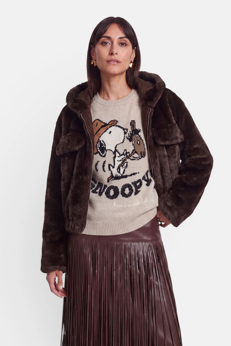 Eine Frau mit glattem braunem Haar trägt eine dunkelbraune Kunstfelljacke mit einem dezenten Reißverschluss, einen beigen Pullover mit einem Snoopy- und Woodstock-Motiv sowie einen braunen Lederrock mit Falten. Sie steht vor einem schlichten weißen Hintergrund und blickt mit ausdruckslosem Gesicht nach vorne.