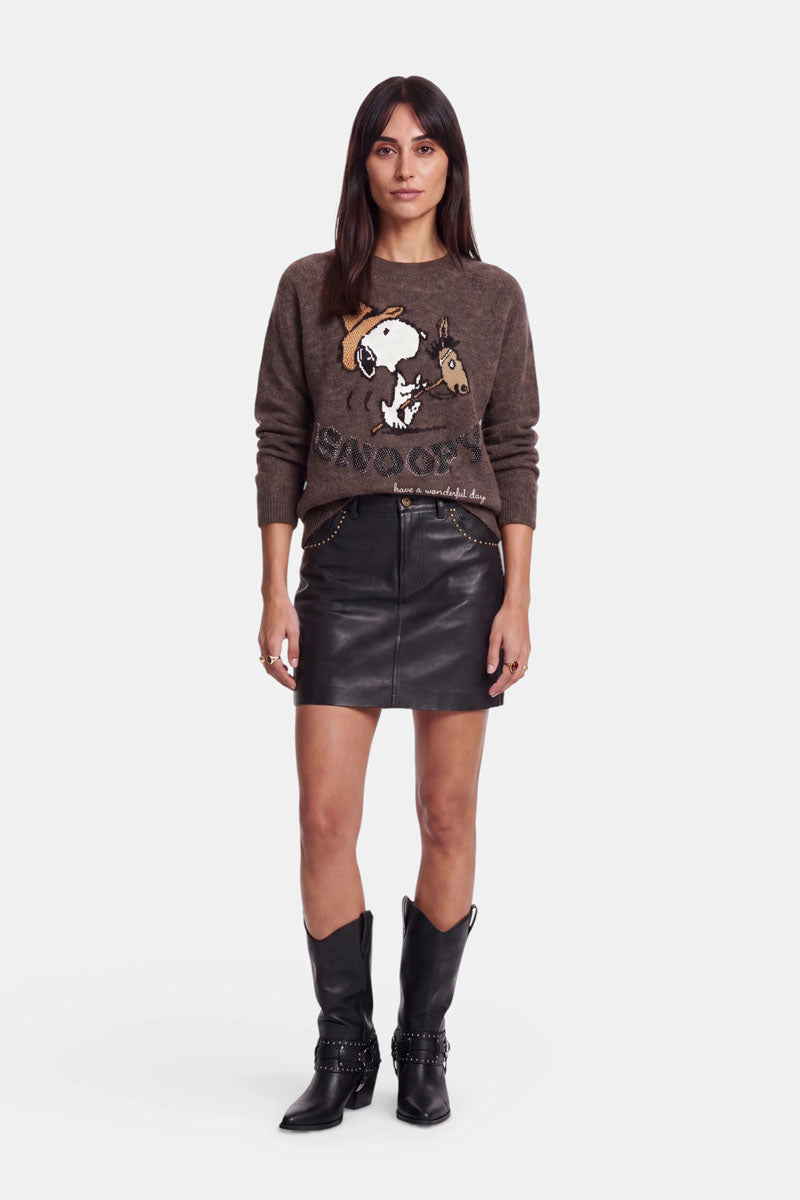 Eine Frau steht mit dem Blick nach vorne und trägt einen braunen Snoopy-Pullover, einen schwarzen Minirock aus veganem Leder mit Nietenverzierung und Reißverschlusstaschen sowie schwarze Cowboystiefel. Sie hat glattes dunkles Haar, hält die Arme entspannt und zeigt einen neutralen Gesichtsausdruck vor einem schlichten weißen Hintergrund.