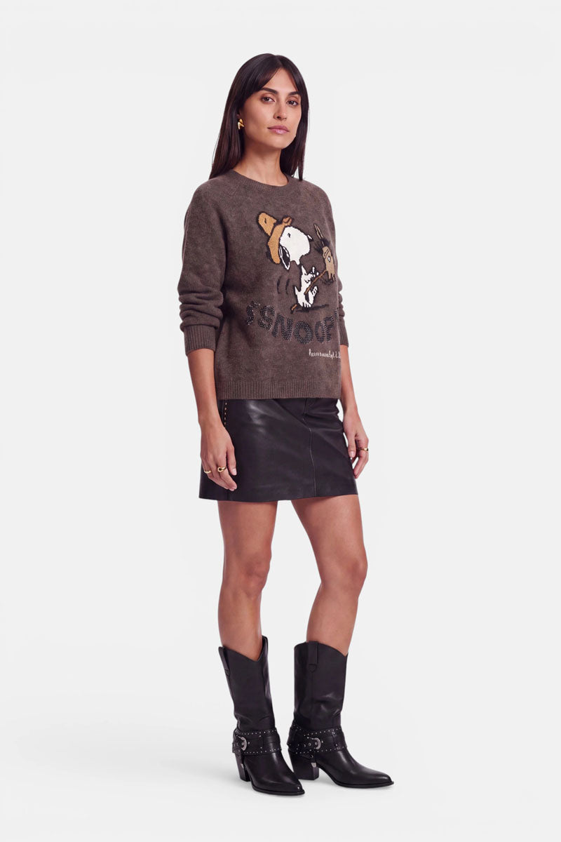 Eine Frau steht vor einem weißen Hintergrund und trägt einen braunen Snoopy-Intarsia-Pullover aus weicher Alpaka-Wolle, dazu einen kurzen schwarzen Lederrock und schwarze Cowboystiefel. Sie hat glattes dunkles Haar und trägt goldene Creolen; ihr Blick ist leicht nach links gerichtet, ihr Gesichtsausdruck ist neutral.