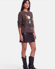 Eine Frau steht vor einem weißen Hintergrund und trägt einen braunen Snoopy-Intarsia-Pullover aus weicher Alpaka-Wolle, dazu einen kurzen schwarzen Lederrock und schwarze Cowboystiefel. Sie hat glattes dunkles Haar und trägt goldene Creolen; ihr Blick ist leicht nach links gerichtet, ihr Gesichtsausdruck ist neutral.