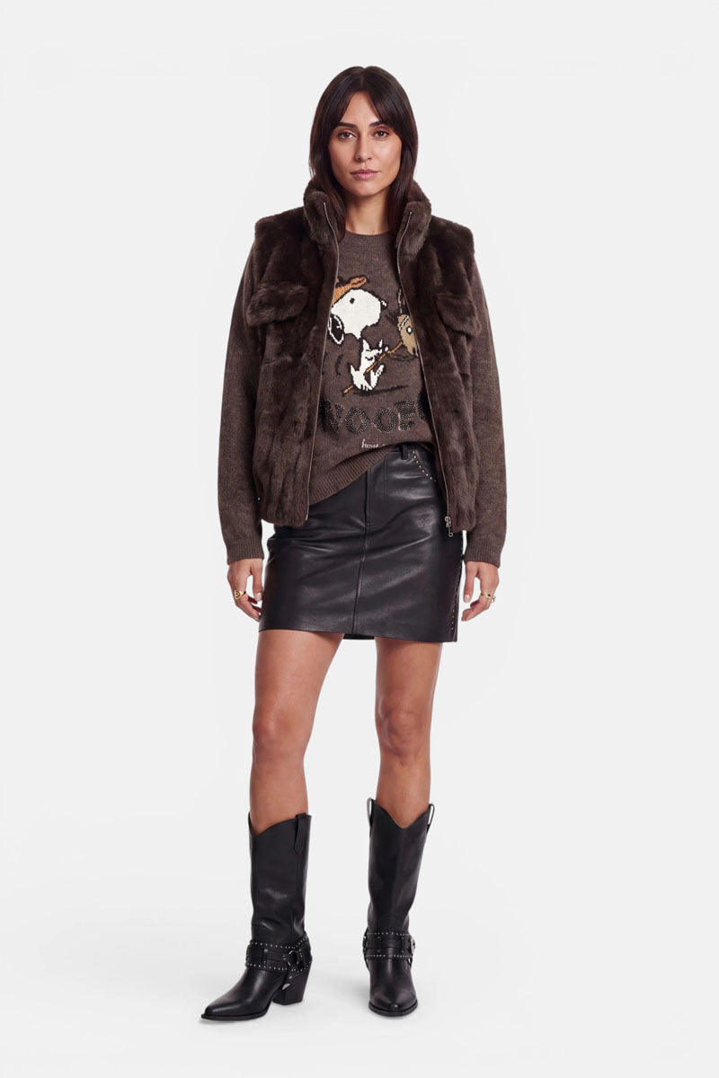 Eine Frau steht mit dem Blick nach vorne vor einem schlichten weißen Hintergrund und trägt eine braune Kunstfellweste mit Stehkragen über einem Pullover mit Snoopy-Motiv, dazu einen schwarzen Lederminirock und schwarze Stiefel im Westernstil mit silbernen Details.