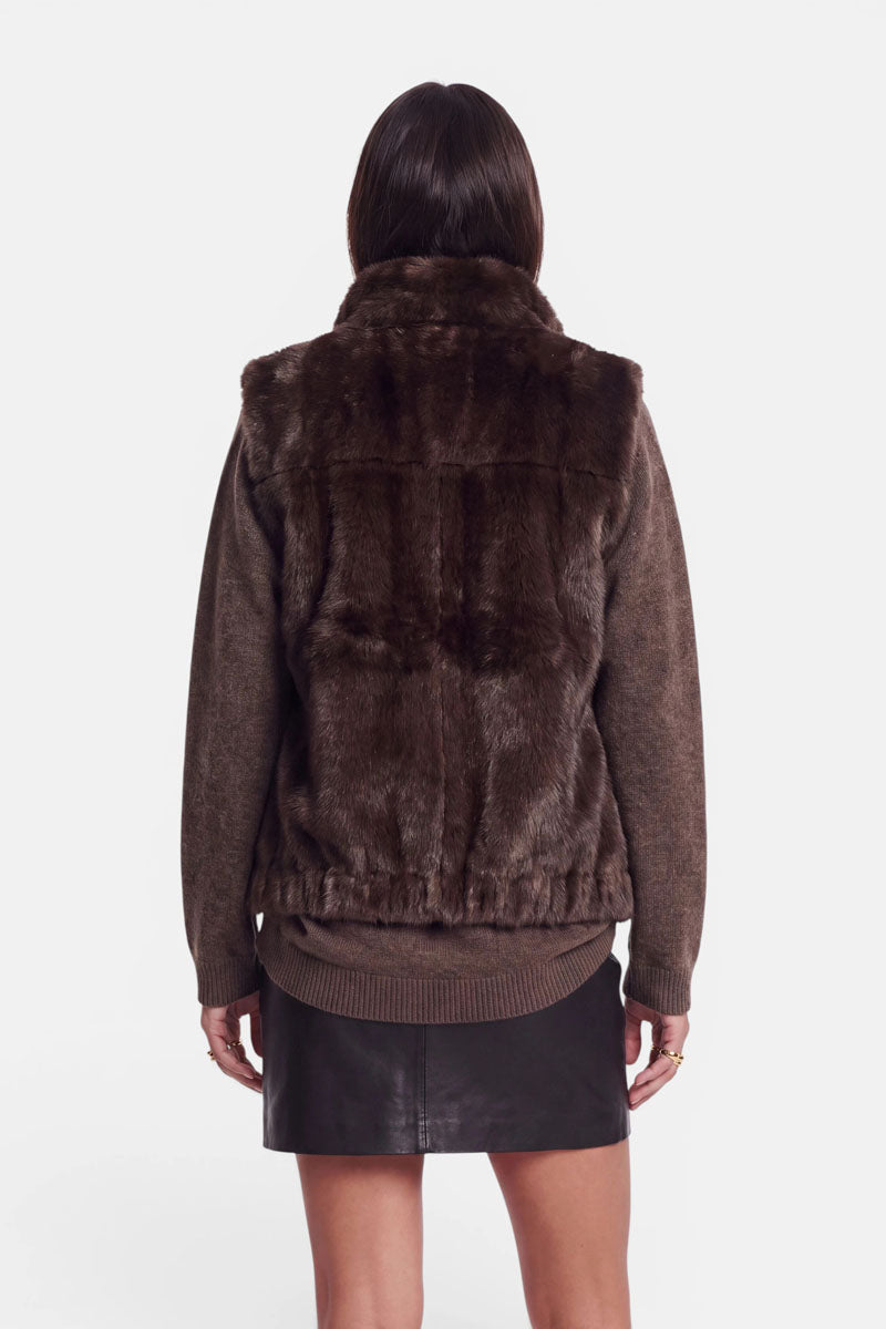 Eine Frau, die mit dem Rücken zur Kamera steht und eine dunkelbraune Kunstfellweste mit Stehkragen über einem braunen Pullover trägt. Außerdem trägt sie einen schwarzen Lederminirock. Ihre Arme hängen entspannt an den Seiten herab. Der Hintergrund ist schlicht weiß, wodurch die Texturen und Farben der Kleidung besonders gut zur Geltung kommen.