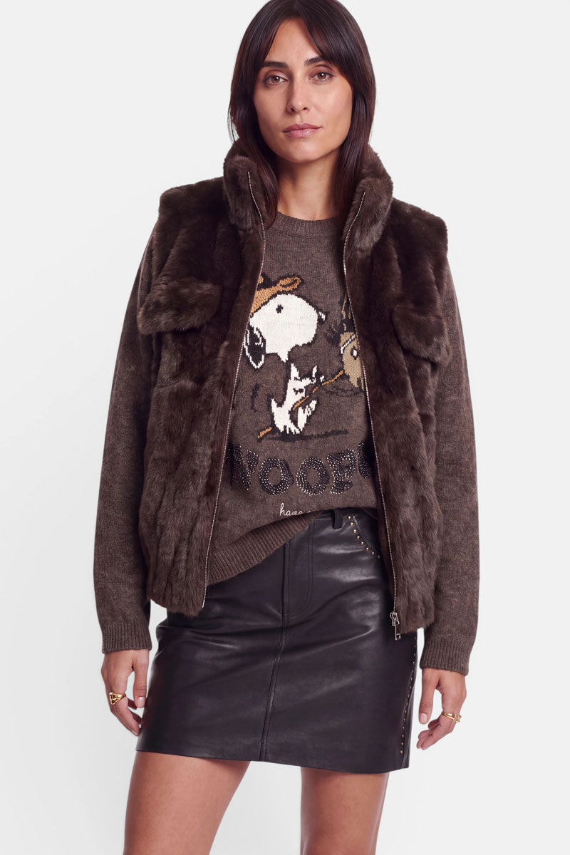 Eine Frau steht in einer braunen Kunstfellweste mit Reißverschluss, die sie über einem Pullover mit Snoopy-Motiv und einem schwarzen Lederrock trägt. Sie hat langes, glattes braunes Haar, ist nur dezent geschminkt und blickt mit ausdruckslosem Gesichtsausdruck vor sich hin, vor einem schlichten weißen Hintergrund.