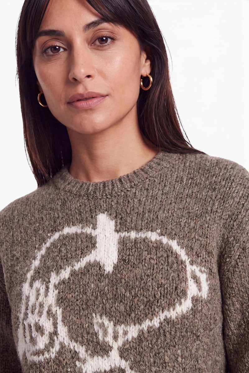 Eine Frau, die einen braunen, grob gestrickten Pullover mit Rundhalsausschnitt trägt, auf dessen Brust ein großes weißes Snoopy-Intarsienmotiv zu sehen ist.