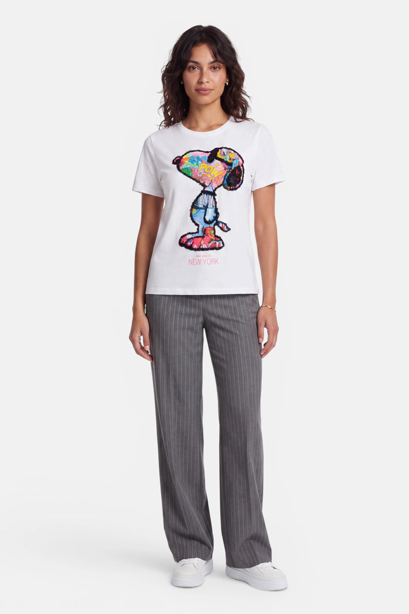 Eine Frau mit welligem braunem Haar trägt ein weißes Baumwolle-Snoopy-T-Shirt mit einem farbenfrohen Pop-Art-Motiv aus Pailletten, eine graue Nadelstreifenhose und weiße Sneaker. Sie steht mit dem Blick nach vorne vor einem schlichten weißen Hintergrund, die Arme entspannt an den Seiten.